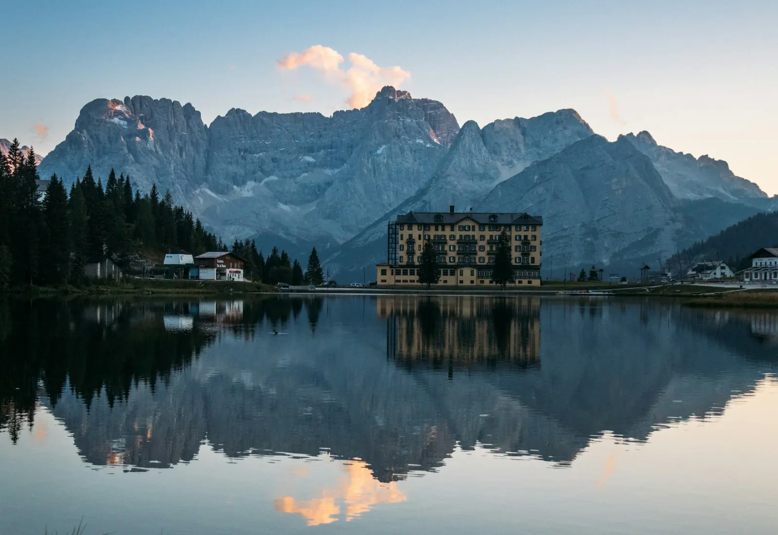 Grand hotel on the shore of an alpine lake, reflected in still water below jagged peaks